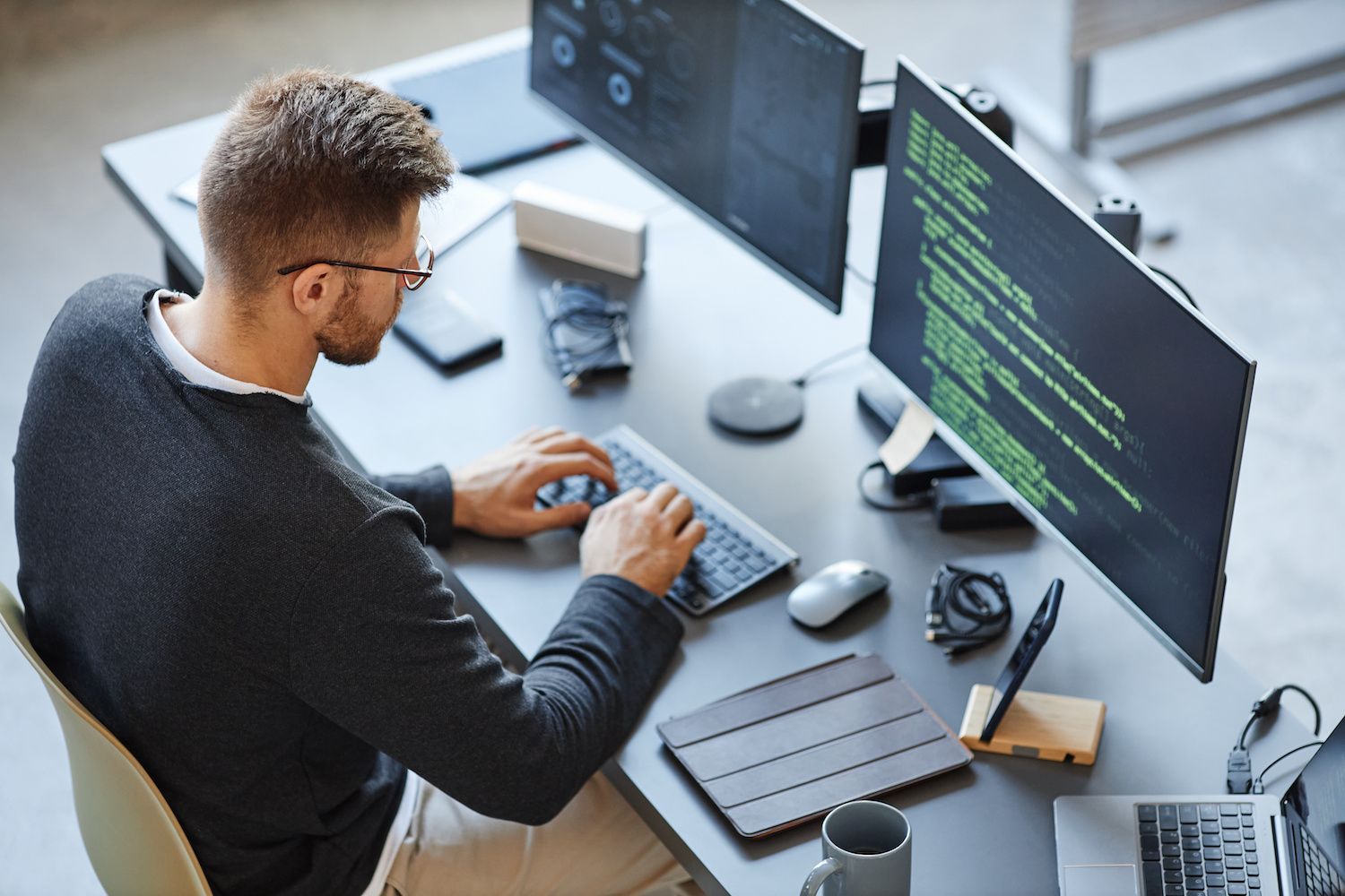 A person is seated at a desk typing on a keyboard while looking at multiple computer monitors displaying lines of code. Various electronic devices and gadgets are placed on the desk. A person is seated at a desk typing on a keyboard while looking at multiple computer monitors displaying lines of code. Various electronic devices and gadgets are placed on the desk.