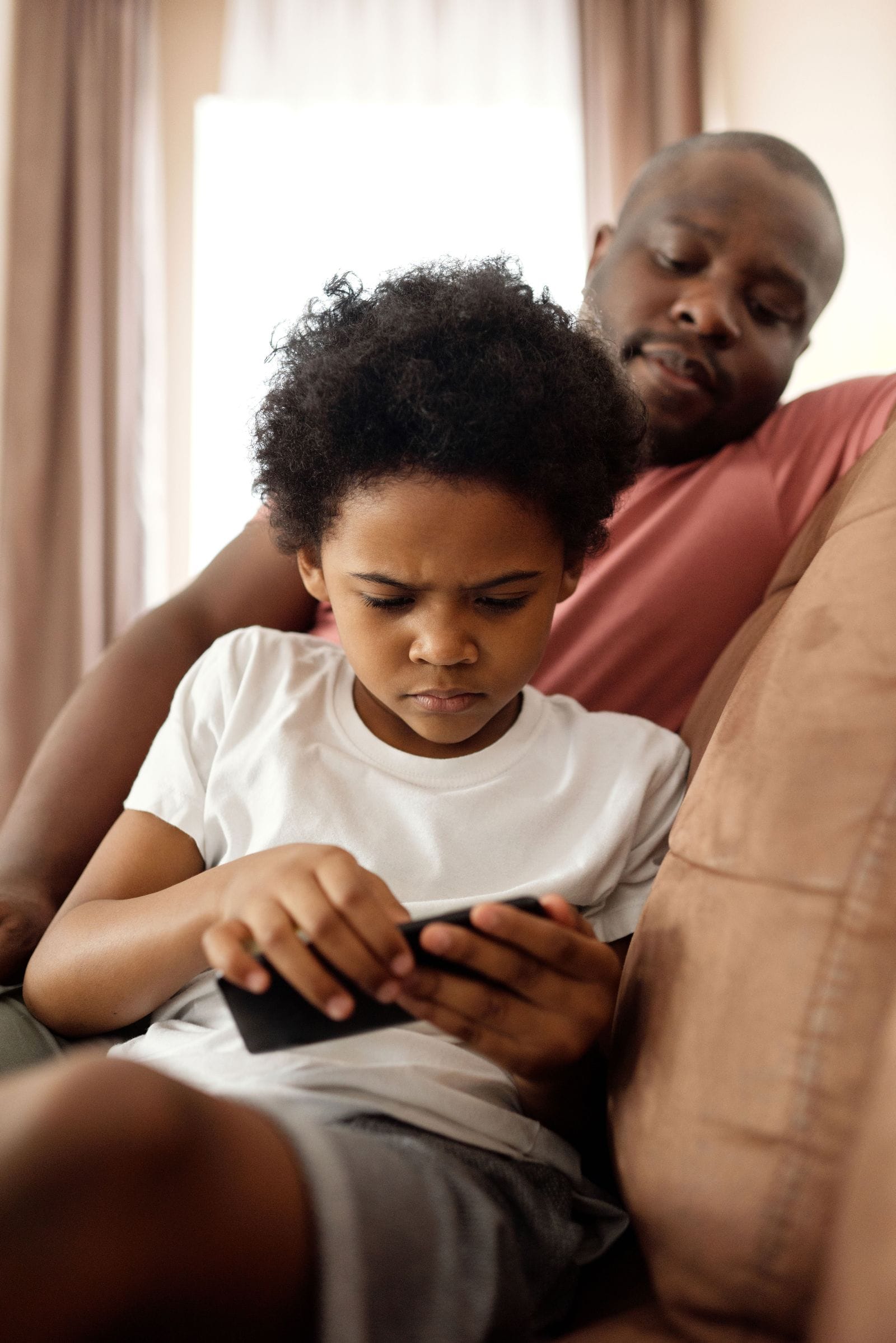 A father looks over his son's shoulder while he uses a smartphone at home, checking device settings together