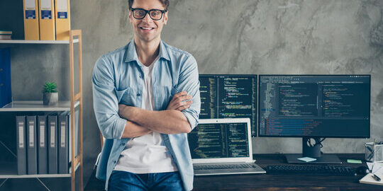 A man stands confidently with arms crossed in front of a desk with two computer monitors displaying code, a laptop, office supplies, and shelves with files and binders.