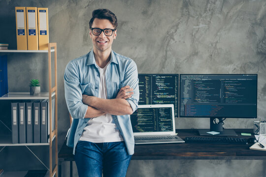 A man stands confidently with arms crossed in front of a desk with two computer monitors displaying code, a laptop, office supplies, and shelves with files and binders. A man stands confidently with arms crossed in front of a desk with two computer monitors displaying code, a laptop, office supplies, and shelves with files and binders.