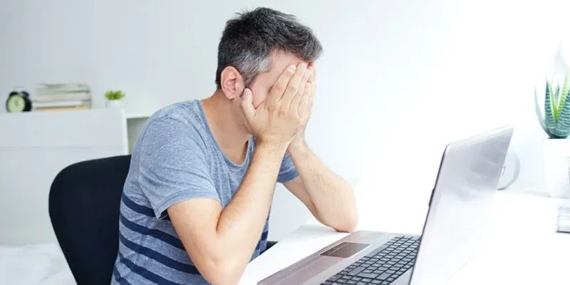 A man in a gray-striped shirt sits at a desk with his head in his hands, facing a laptop in a bright, tidy room. A man in a gray-striped shirt sits at a desk with his head in his hands, facing a laptop in a bright, tidy room.