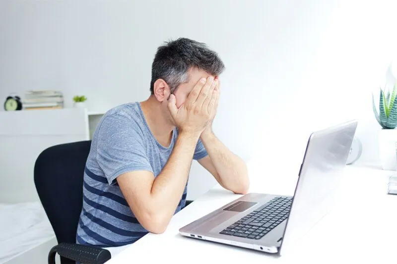 A man in a gray-striped shirt sits at a desk with his head in his hands, facing a laptop in a bright, tidy room. A man in a gray-striped shirt sits at a desk with his head in his hands, facing a laptop in a bright, tidy room.