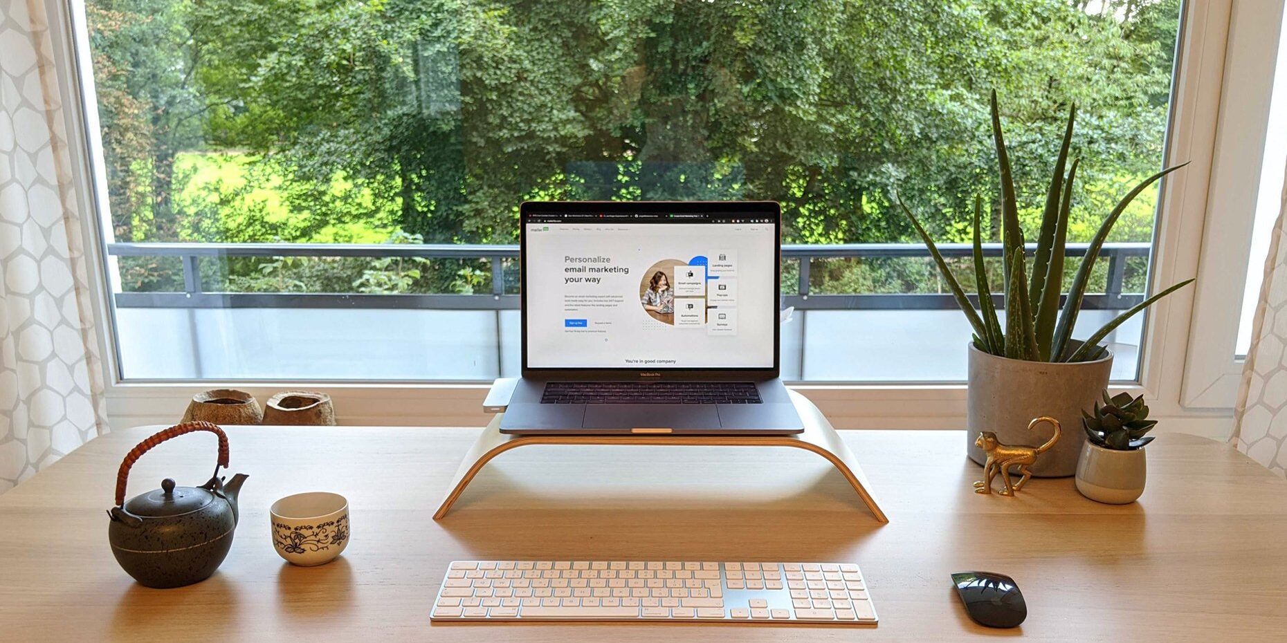 A tidy desk with a laptop on a stand, a keyboard, a mouse, a teapot, a cup, and a potted plant. A large window behind shows trees and greenery outside. A tidy desk with a laptop on a stand, a keyboard, a mouse, a teapot, a cup, and a potted plant. A large window behind shows trees and greenery outside.