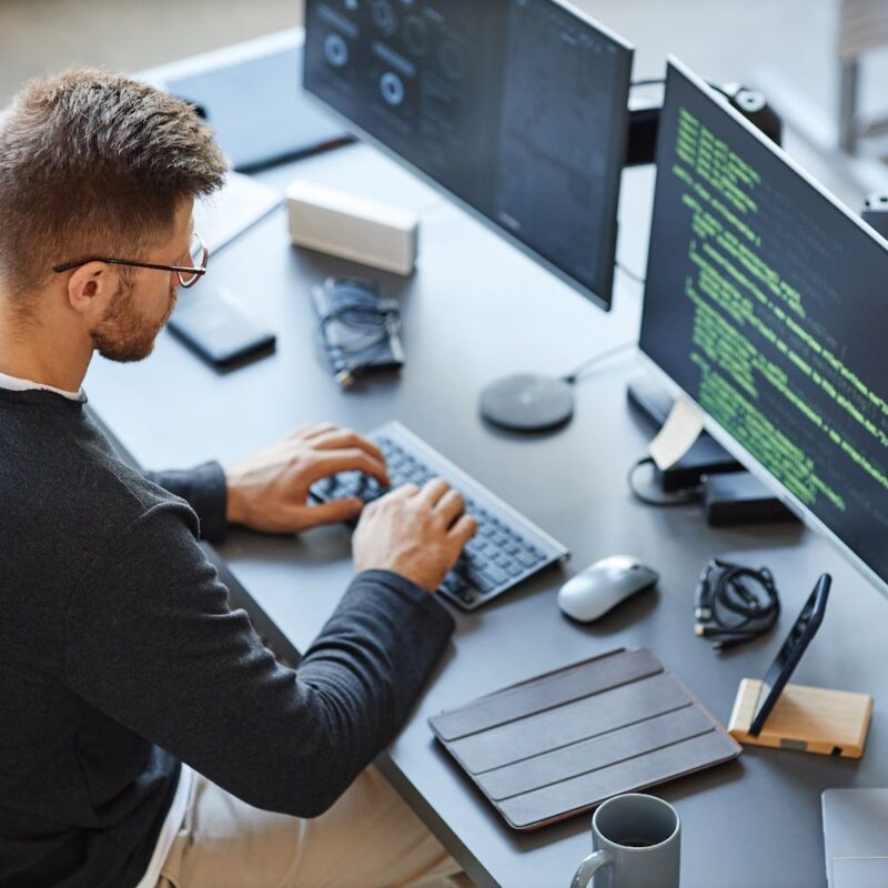 A person is seated at a desk typing on a keyboard while looking at multiple computer monitors displaying lines of code. Various electronic devices and gadgets are placed on the desk.