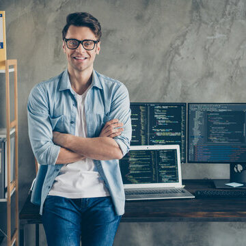 A man stands confidently with arms crossed in front of a desk with two computer monitors displaying code, a laptop, office supplies, and shelves with files and binders.