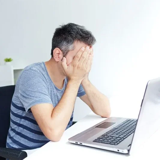 A man in a gray-striped shirt sits at a desk with his head in his hands, facing a laptop in a bright, tidy room.
