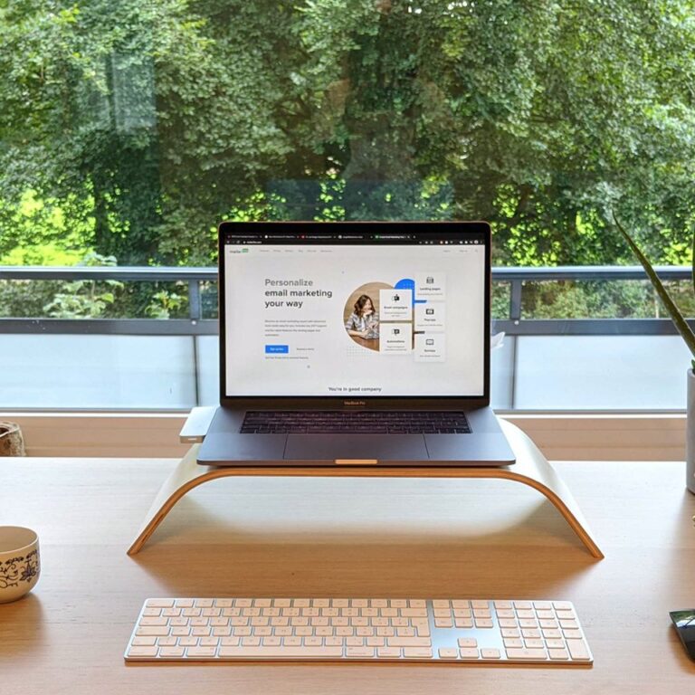 A tidy desk with a laptop on a stand, a keyboard, a mouse, a teapot, a cup, and a potted plant. A large window behind shows trees and greenery outside.