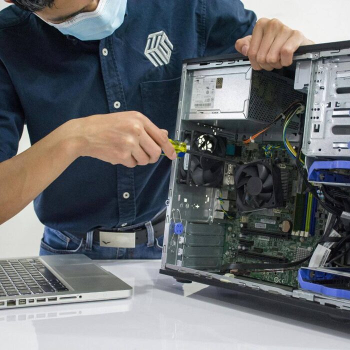 Person wearing a mask repairs an open desktop computer with a screwdriver, next to a closed laptop on a white table.